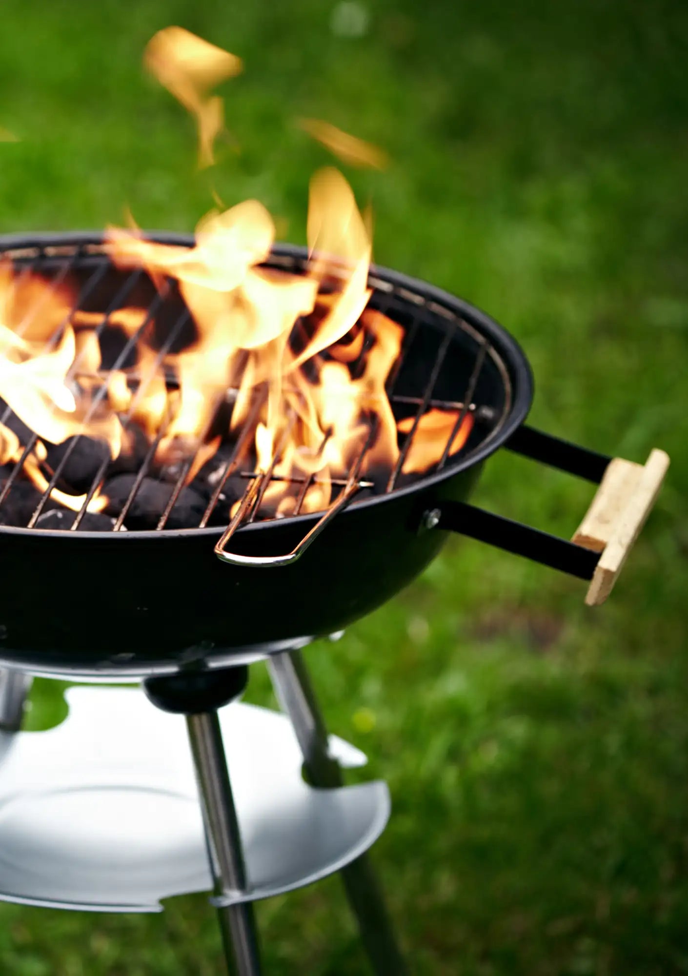Schwarzer Holzkohlengrill mit Holzegriffen und Metallbeinen, Flammen lecken an den Kohlen im Inneren.