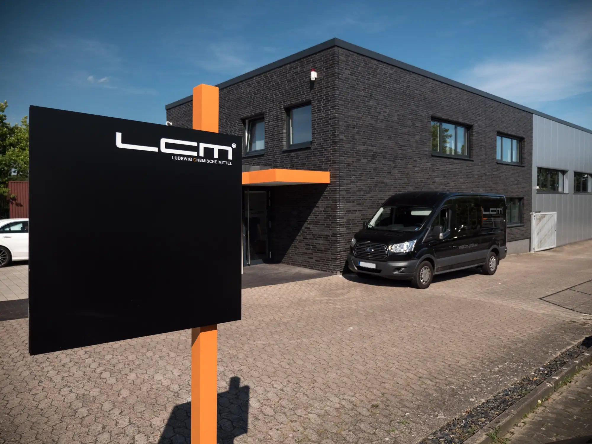 Schwarzes rechteckiges Schild mit weißem ’lcm’-Logo und kleinerem Text darunter, montiert auf einem orangen Pfahl.